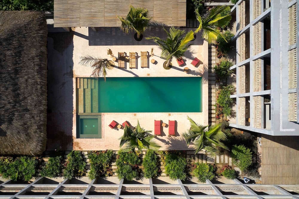 Aerial view of a hotel courtyard featuring a rectangular turquoise pool, lounge chairs, palm trees, and surrounding greenery.